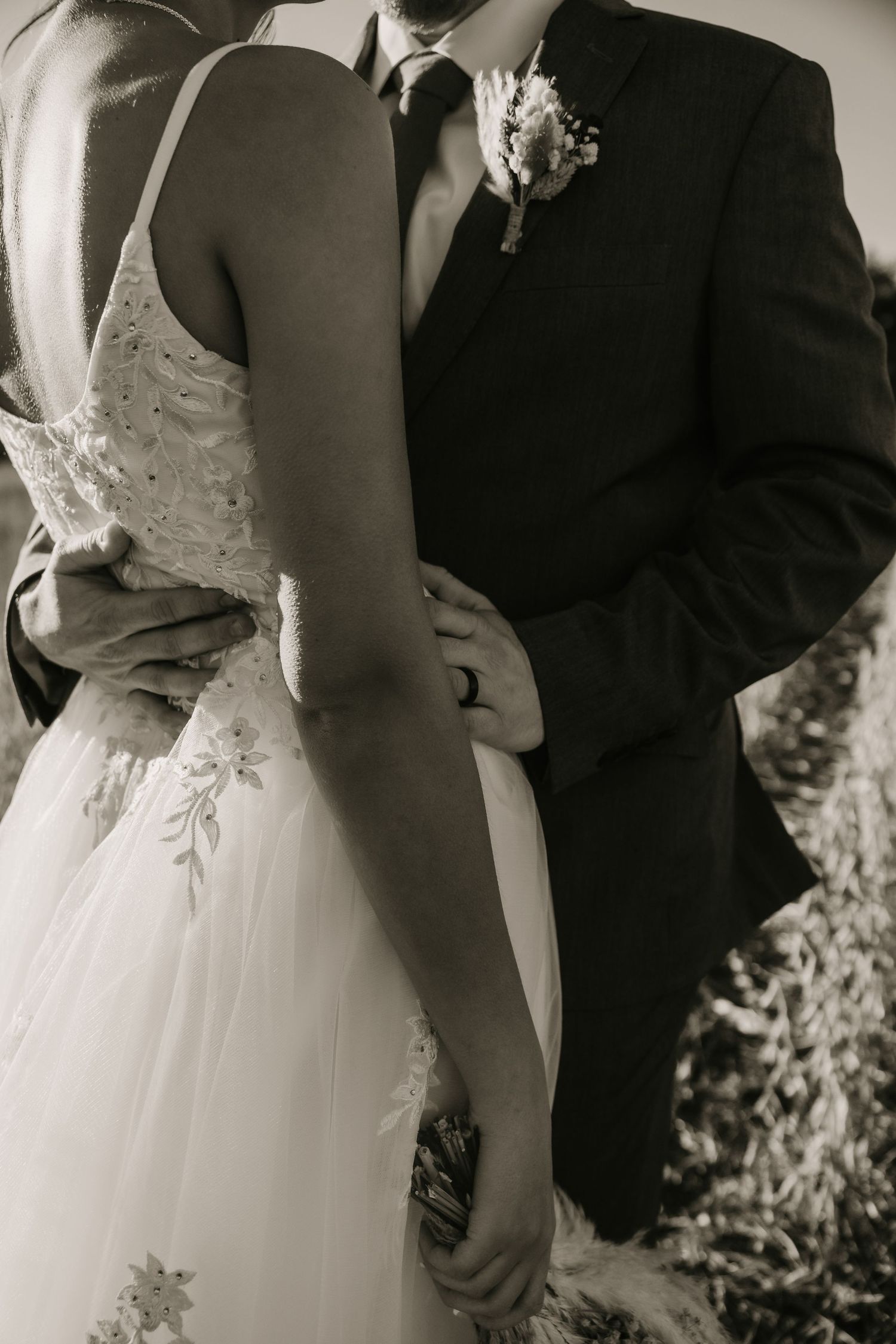 Black and white photo of newlyweds embracing, showing detailed lace dress and groom's boutonniere.