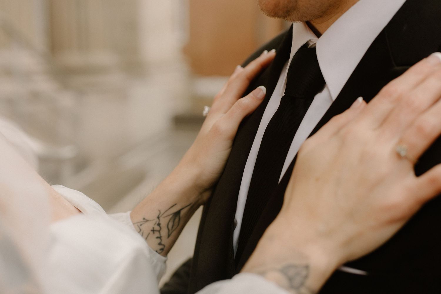 Intimate close-up of tattooed hands against a black suit jacket during a wedding moment.