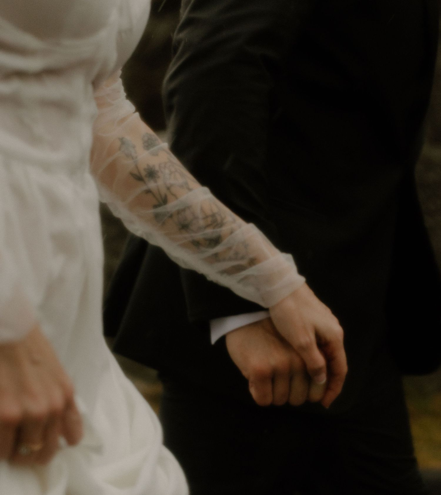 A tattooed arm in white reaching to hold hands with someone in black clothing.