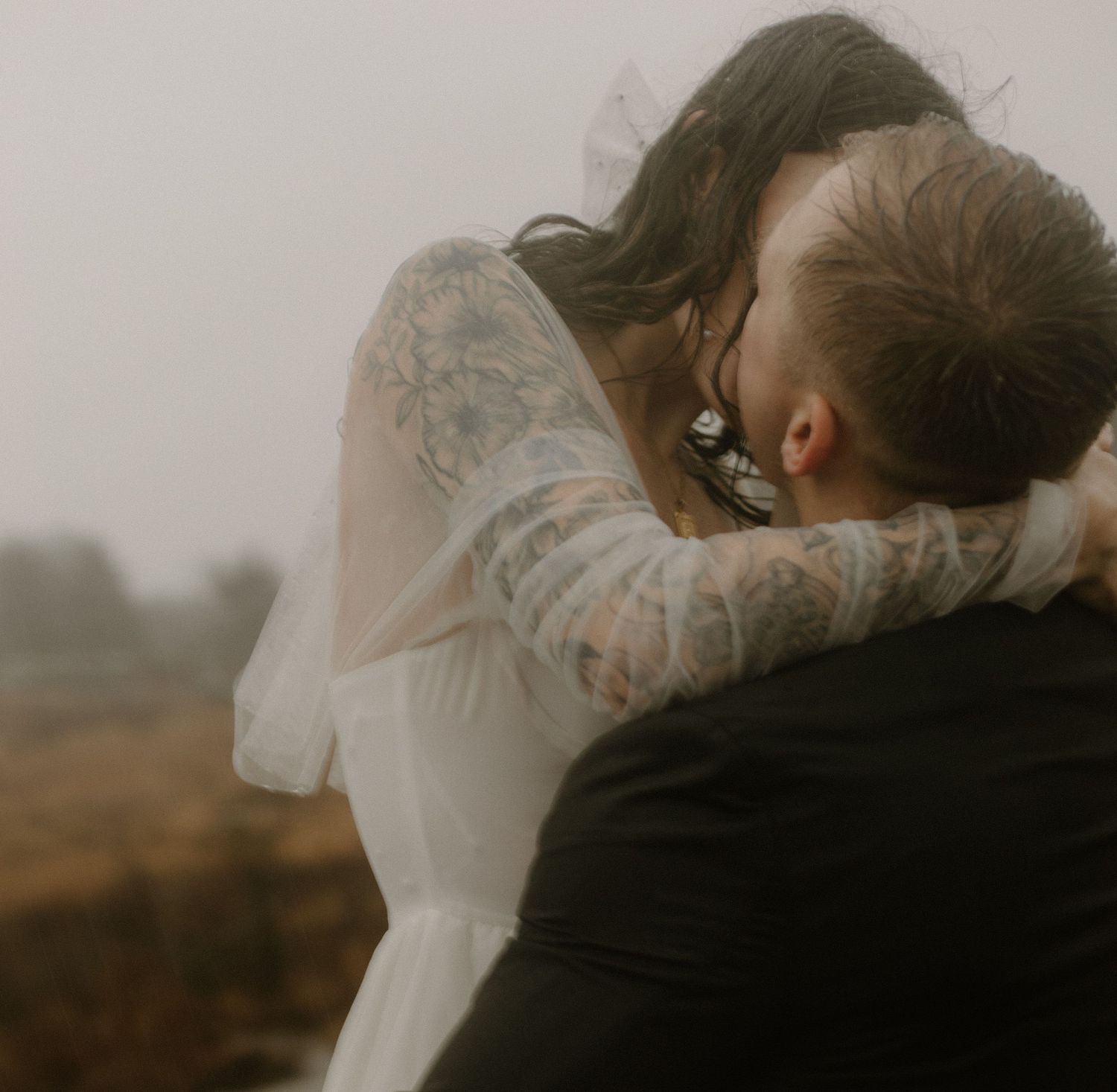 An intimate wedding moment captured in muted tones as a couple shares a tender embrace against a misty backdrop.