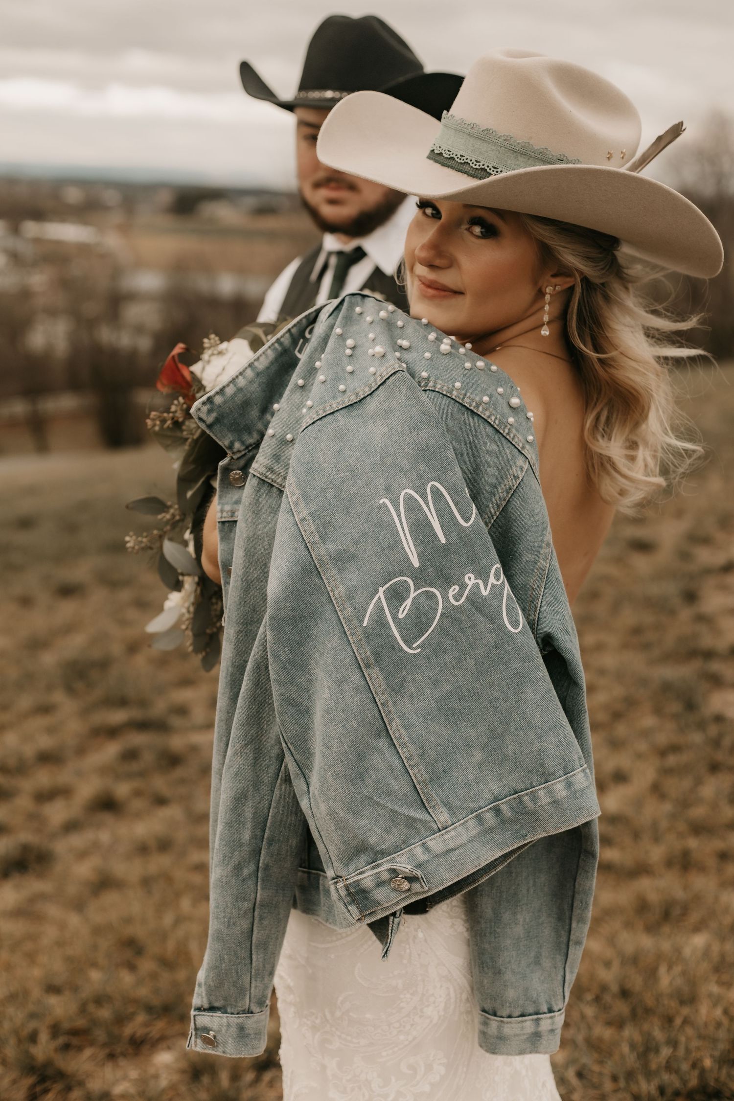 Bride wearing customized denim jacket over wedding dress poses with groom in western style.