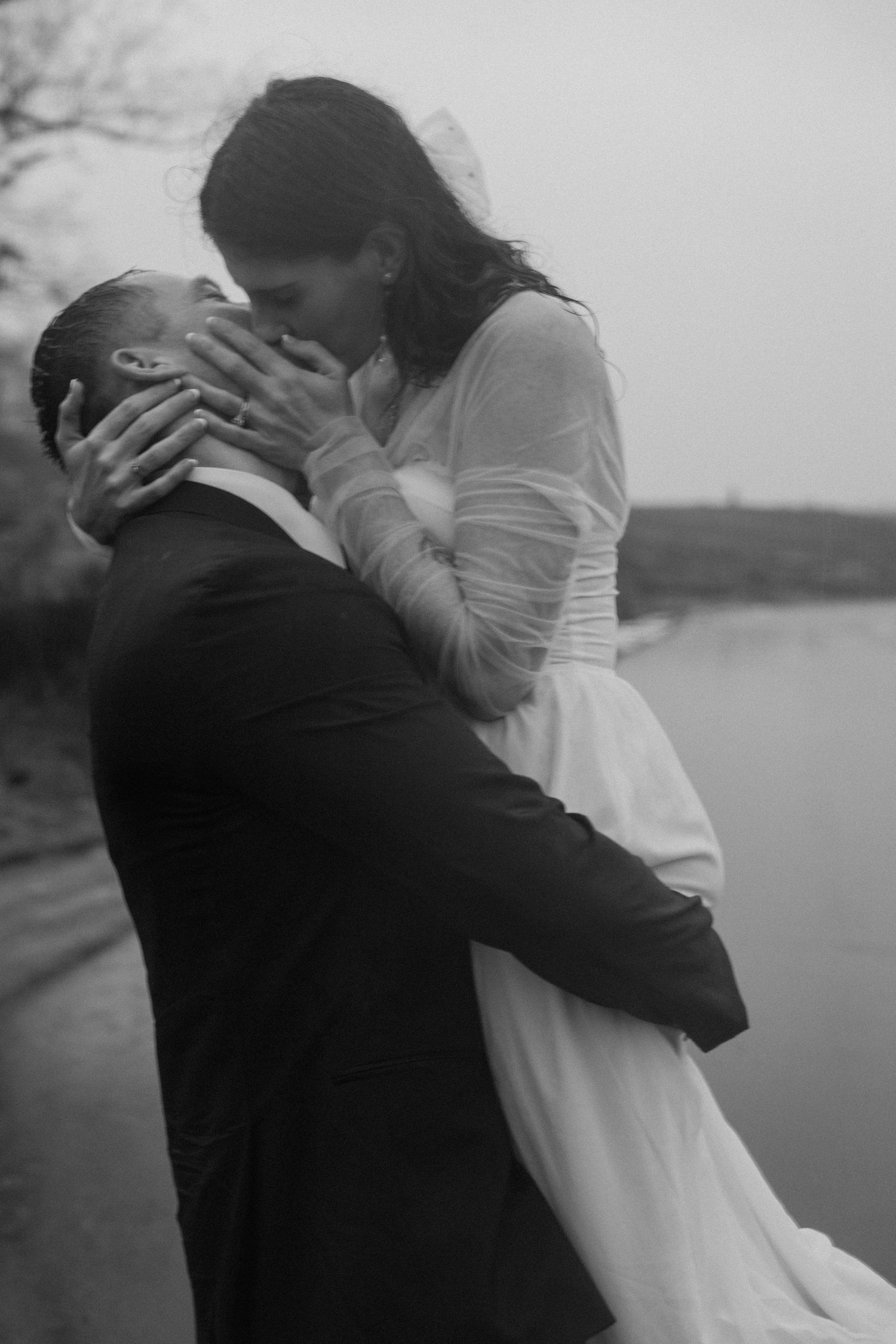 A romantic black and white photo of a couple embracing by a lake, with the woman in white leaning down to kiss her partner.