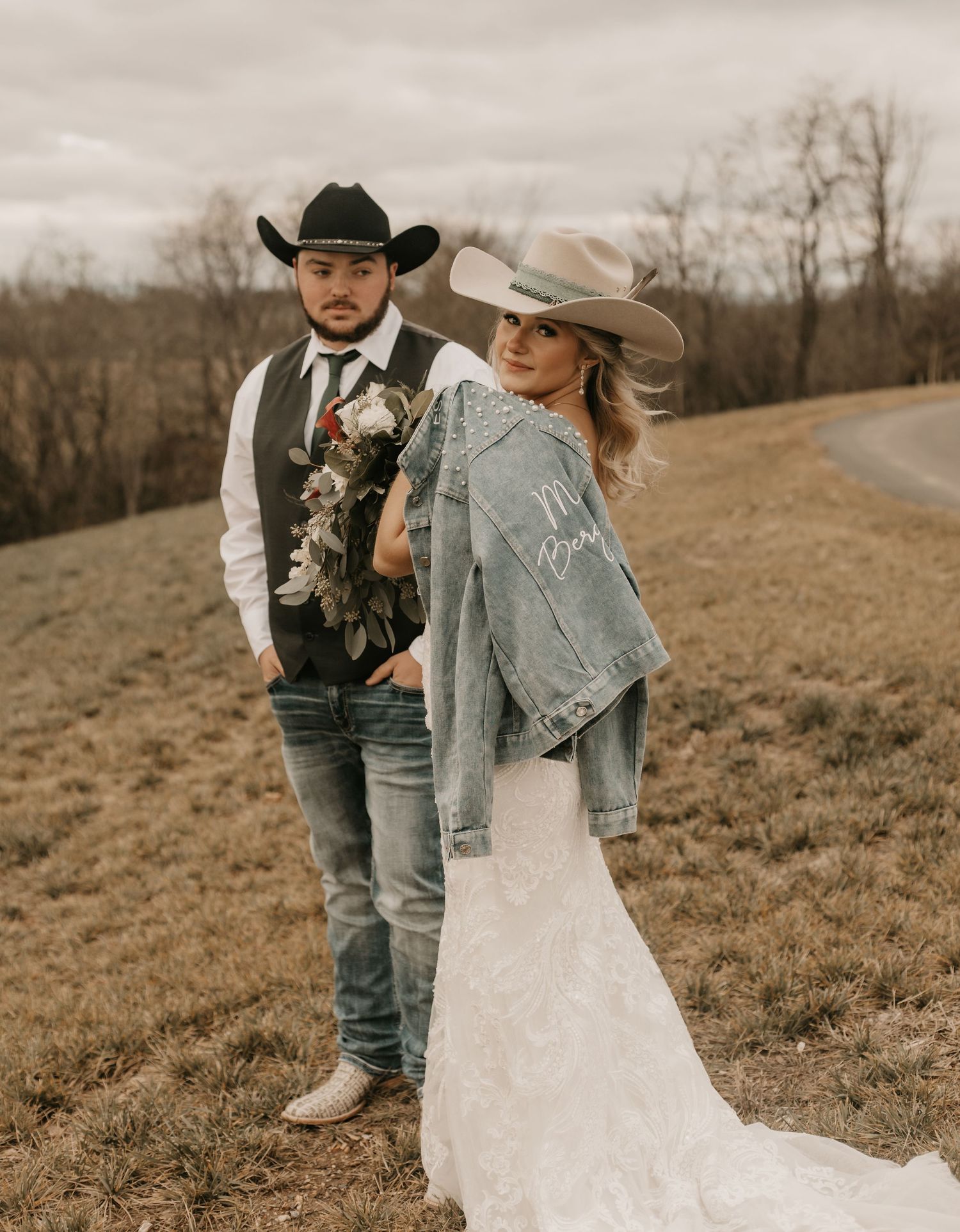 Couple in western attire pose for wedding photos on a hillside with rustic scenery.