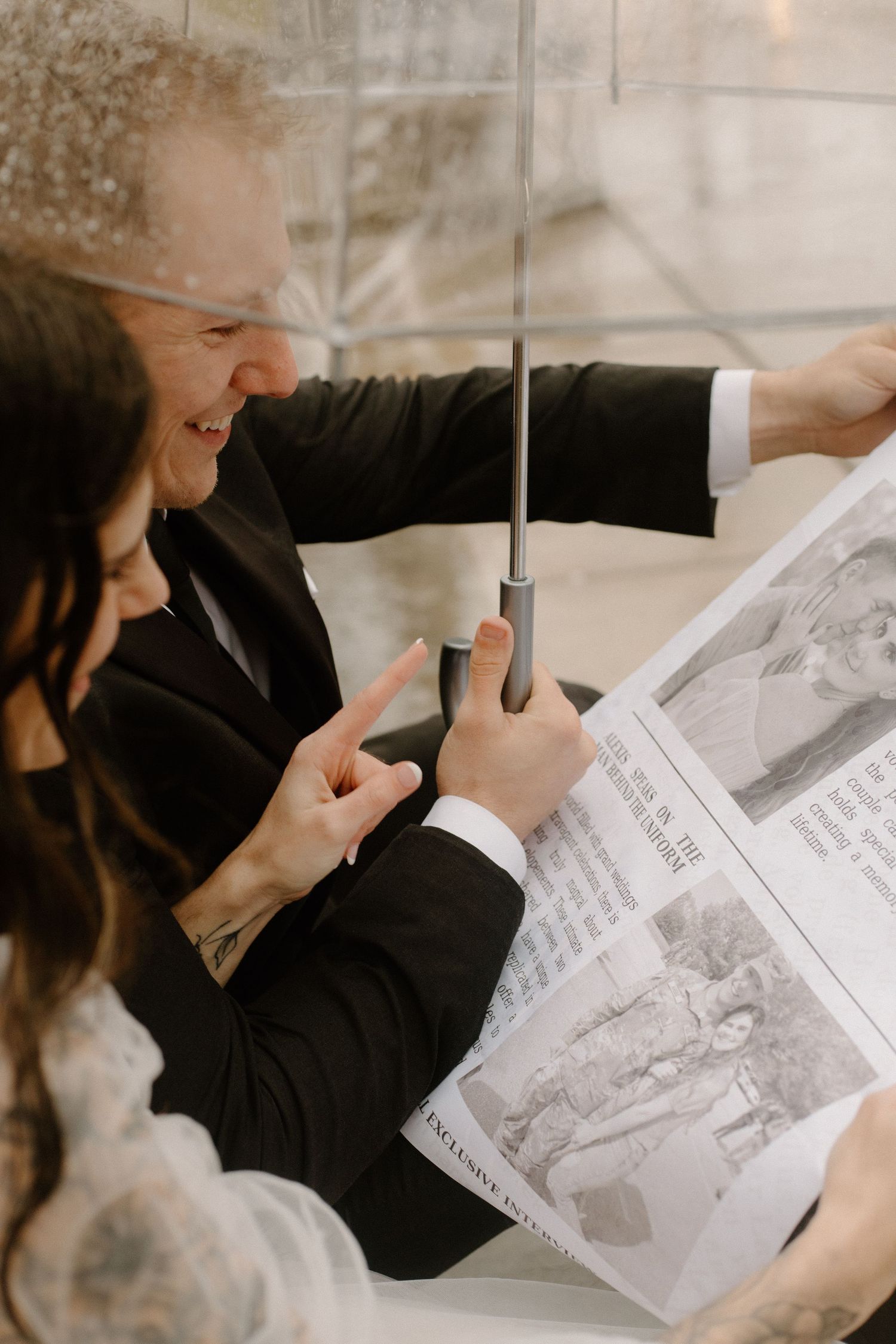A couple sharing laughter while reading a newspaper under an umbrella.