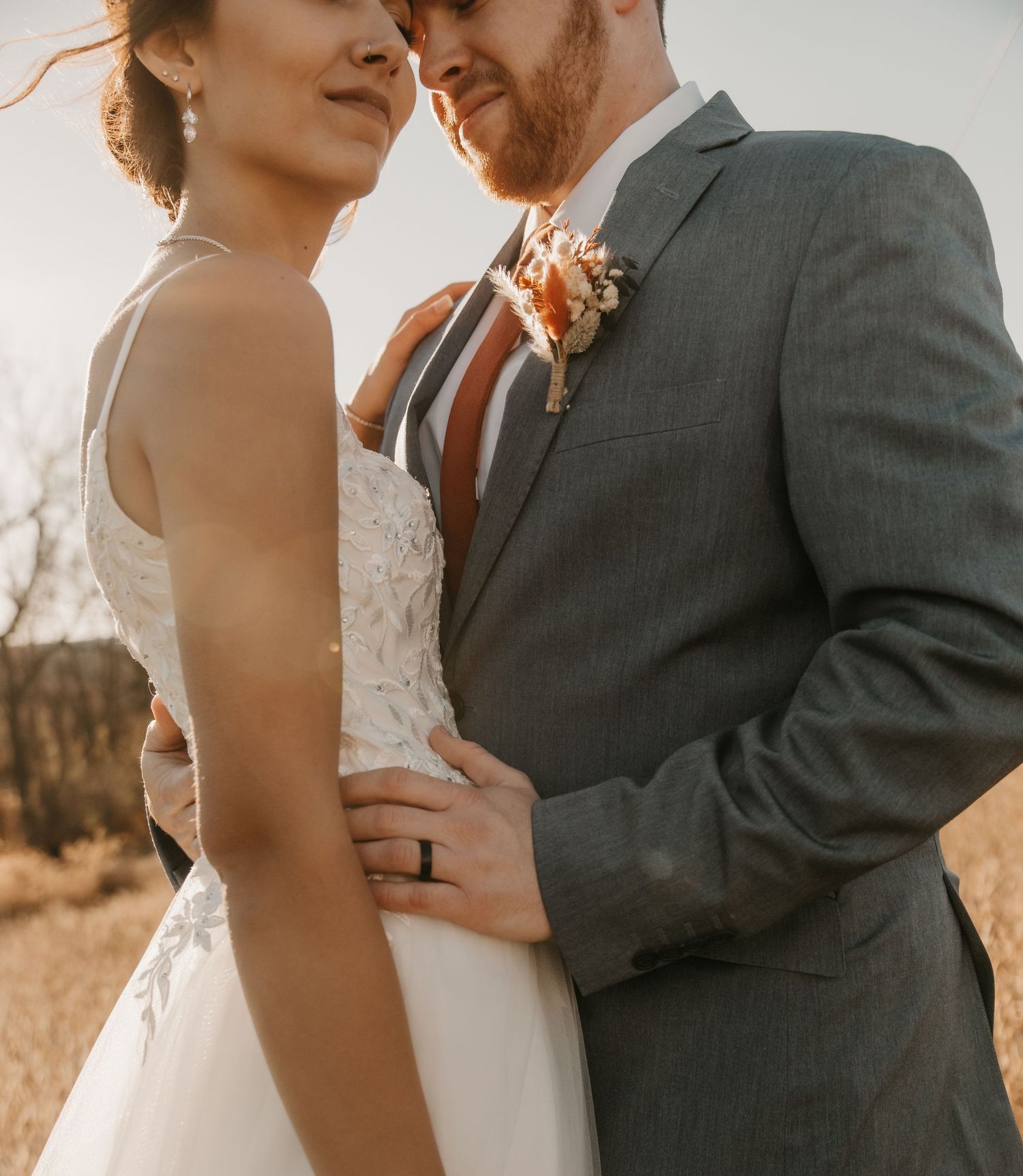 Sun-drenched wedding portrait showing intricate dress details and groom's gray suit.