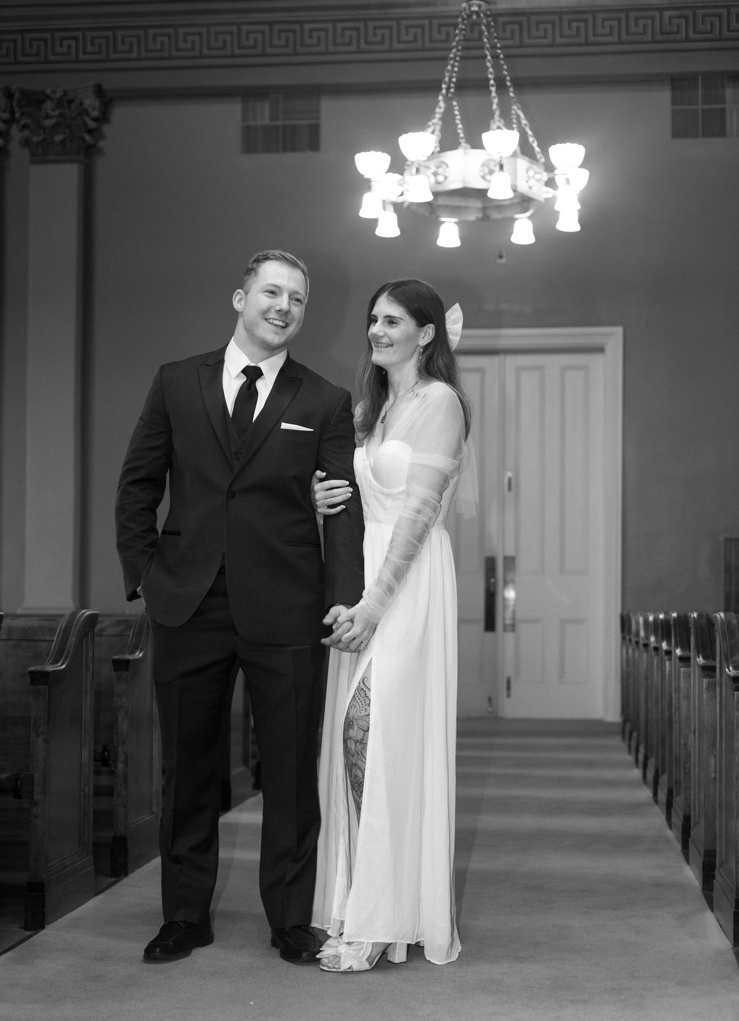 A formal couple standing in a church aisle under a chandelier.