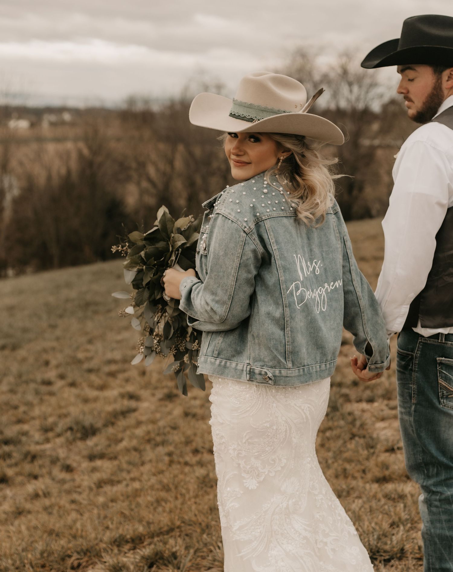 A western-style wedding photo featuring a denim jacket with text and white dress against a rural landscape.