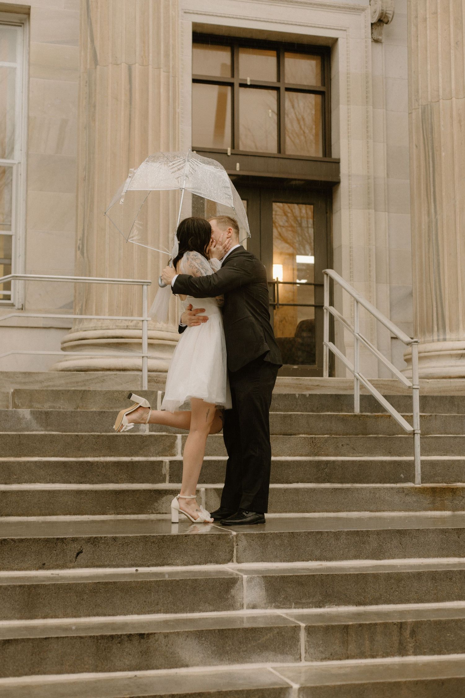 A couple embraces on stone steps with a clear umbrella outside a building.