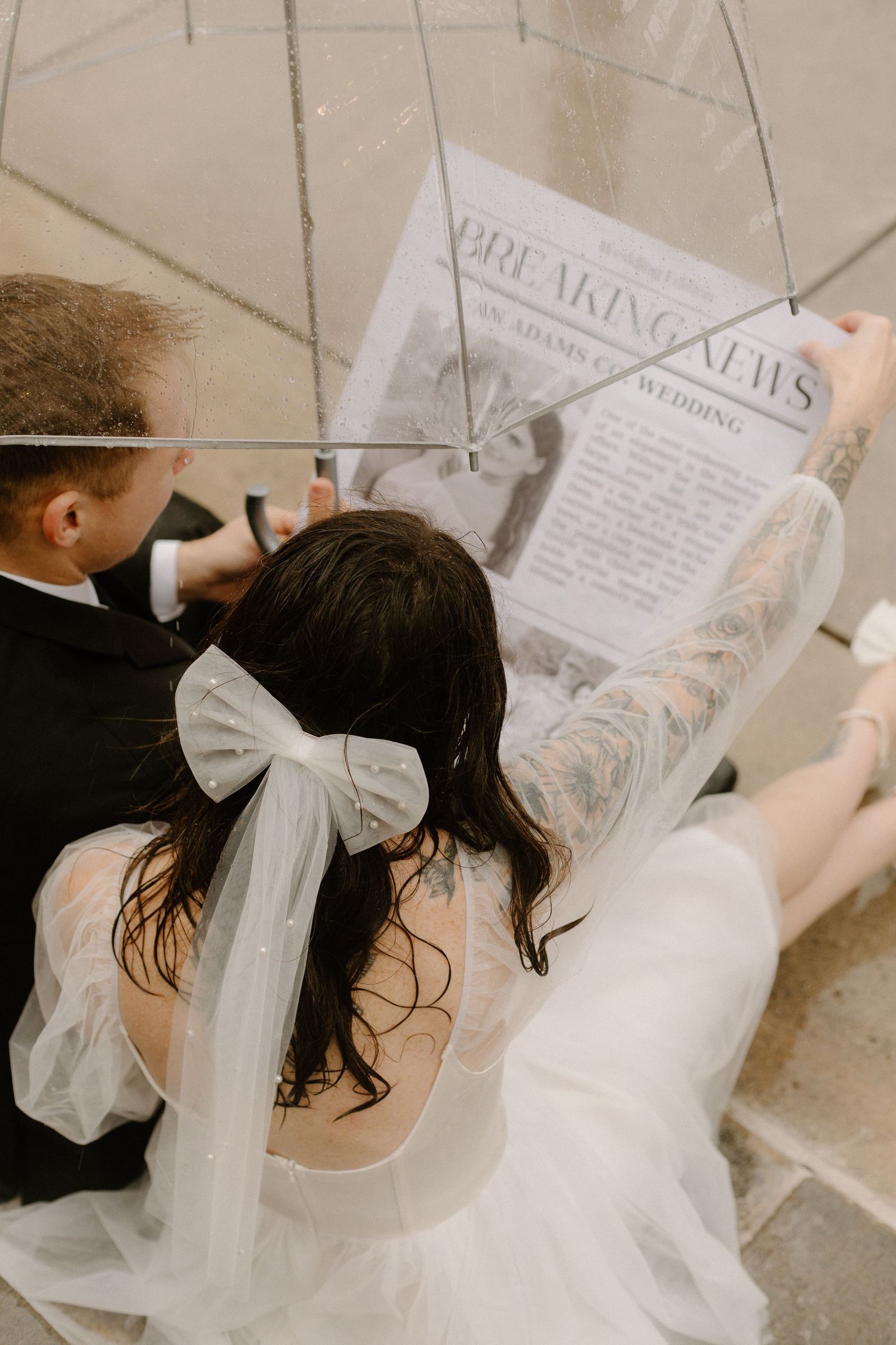 A couple in wedding attire reading a newspaper together, with a white bow detail visible on the bride's dress.
