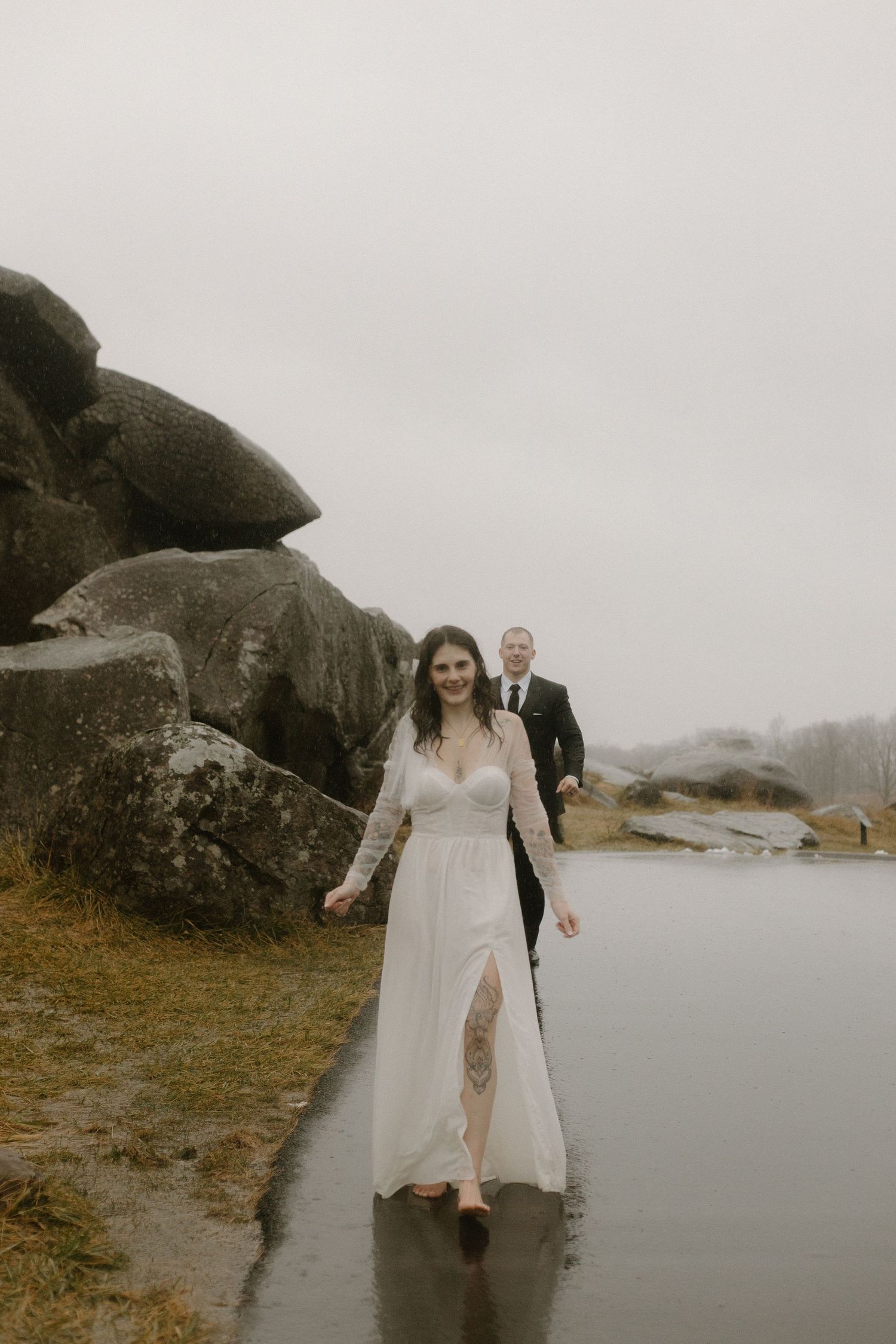 A bride in a flowing white dress walks along a wet path near large rock formations.