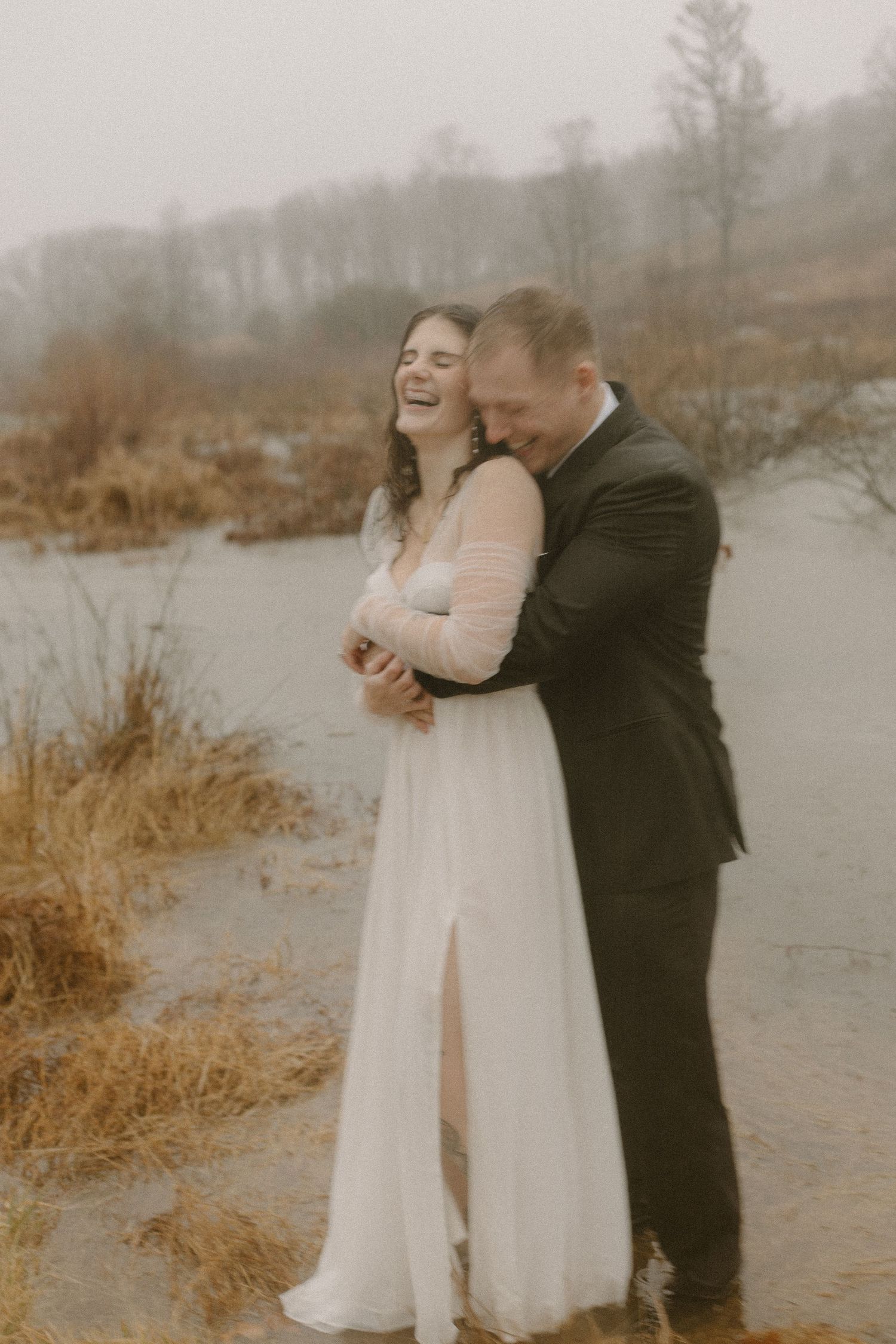 Joyful couple embracing and laughing in wedding attire by a lake.