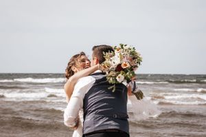 Newlyweds embracing on windy beach with bride holding floral bouquet.