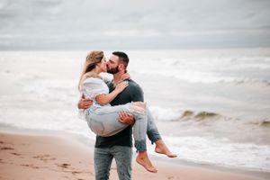 Couple embracing on beach with waves in background as partner carries other in romantic pose.