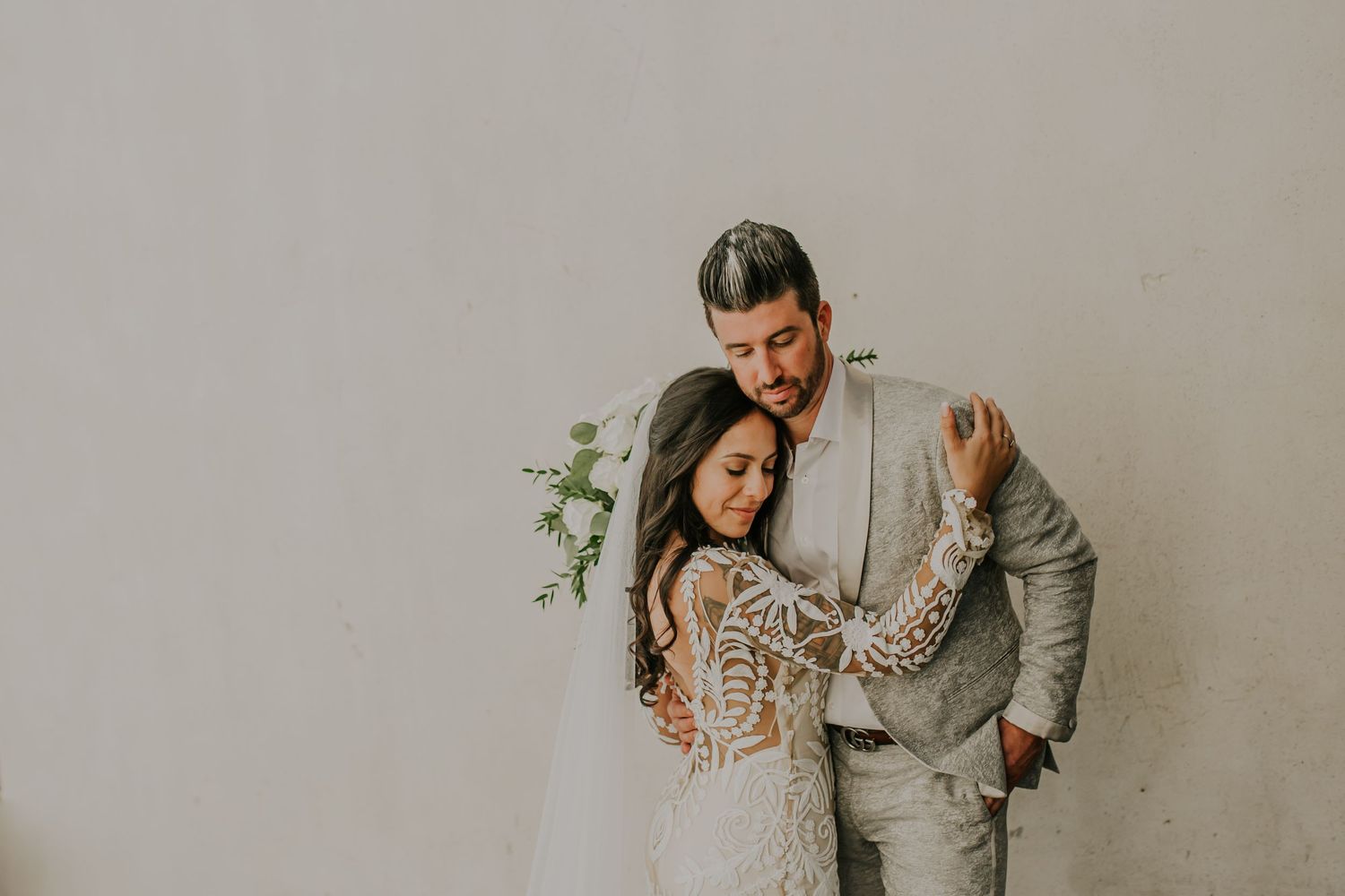A romantic wedding portrait against a cream wall with a lace dress and grey suit on a couple standing close together.