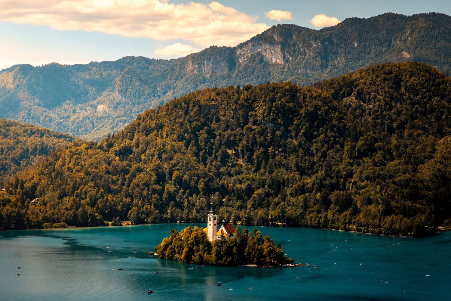 A small church on an island in a turquoise lake surrounded by forested mountains during sunset.