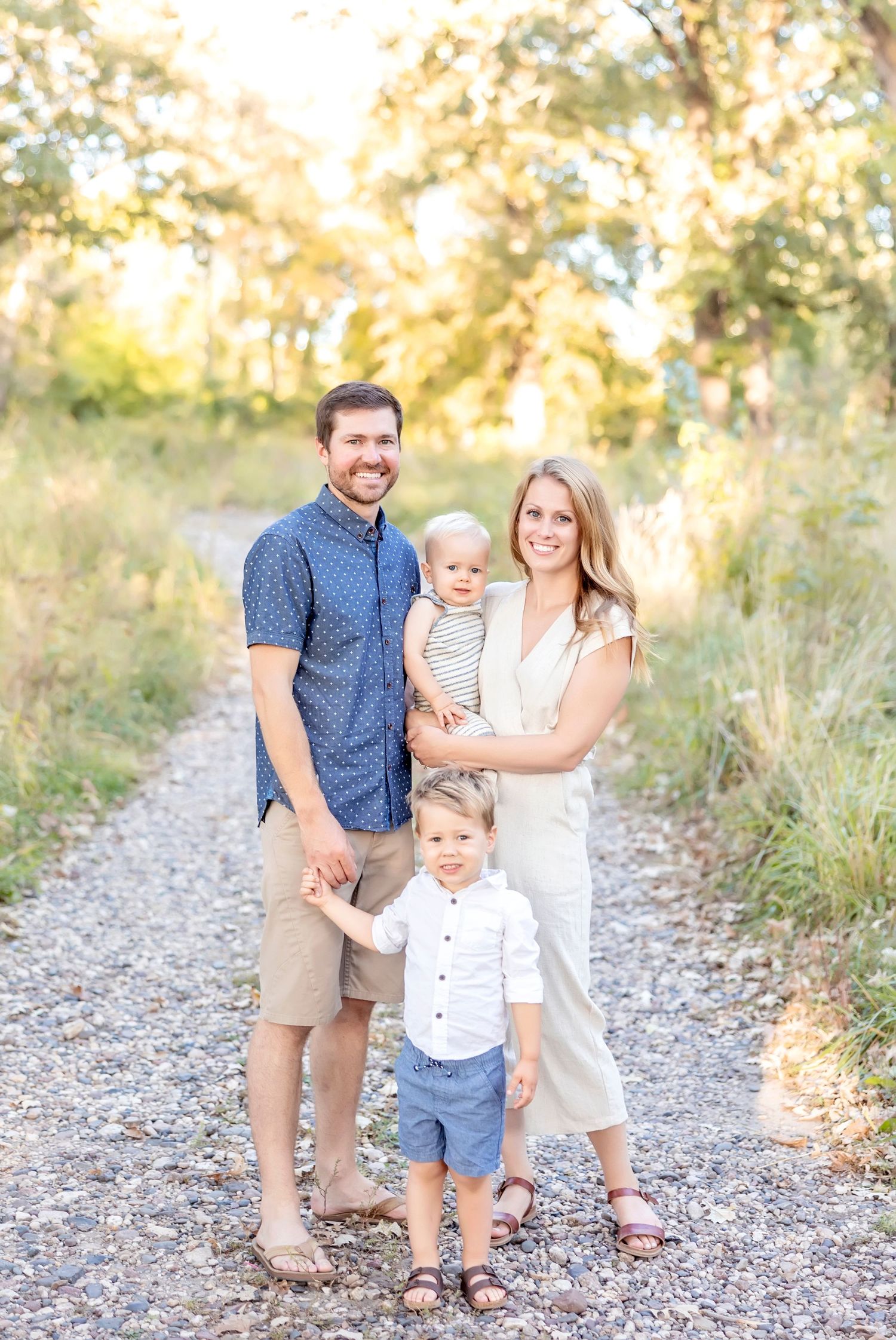 A happy family poses together on a gravel path surrounded by greenery during a summer photo session.
