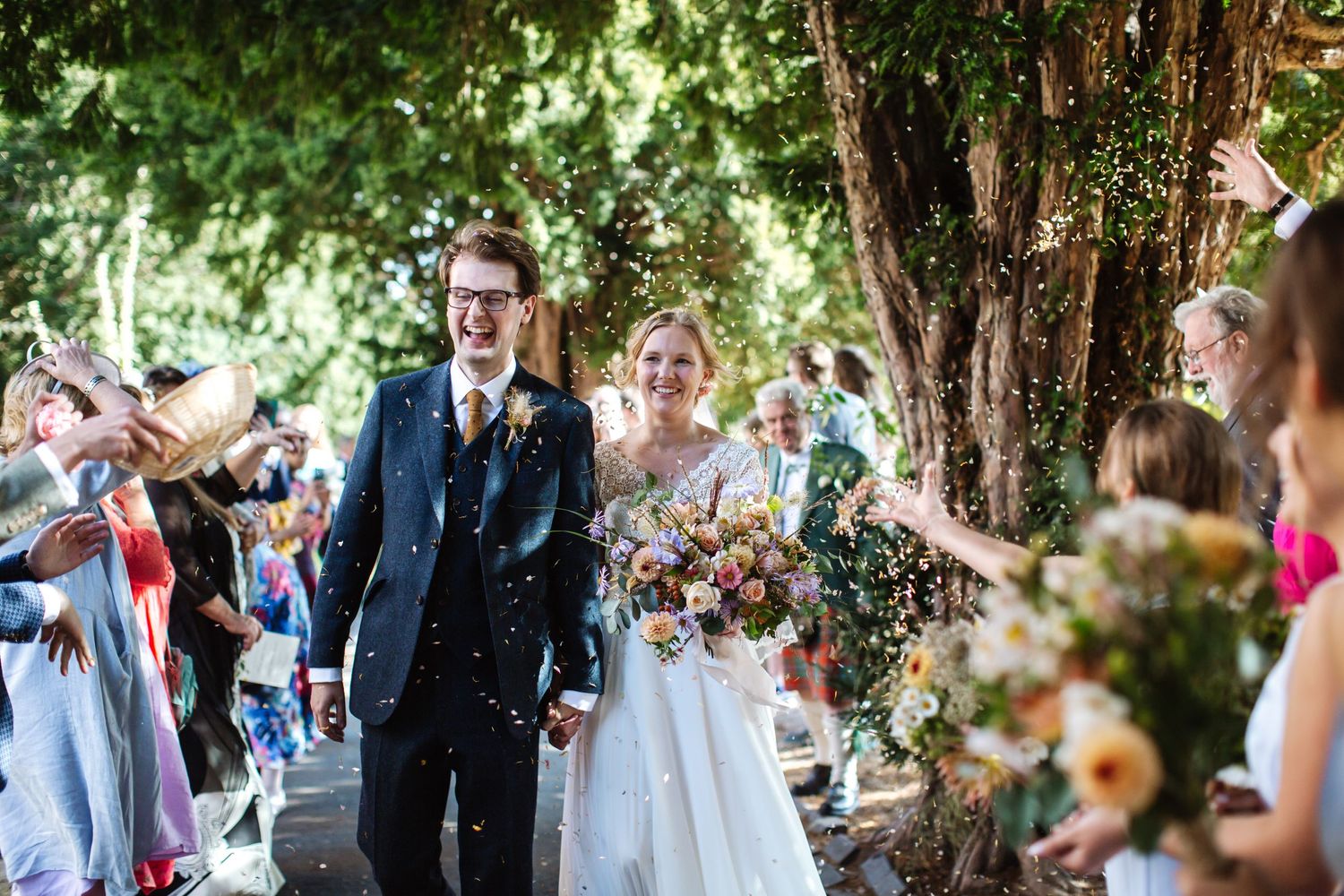 Wedding guests throw colorful confetti over the newlyweds under a tree canopy.