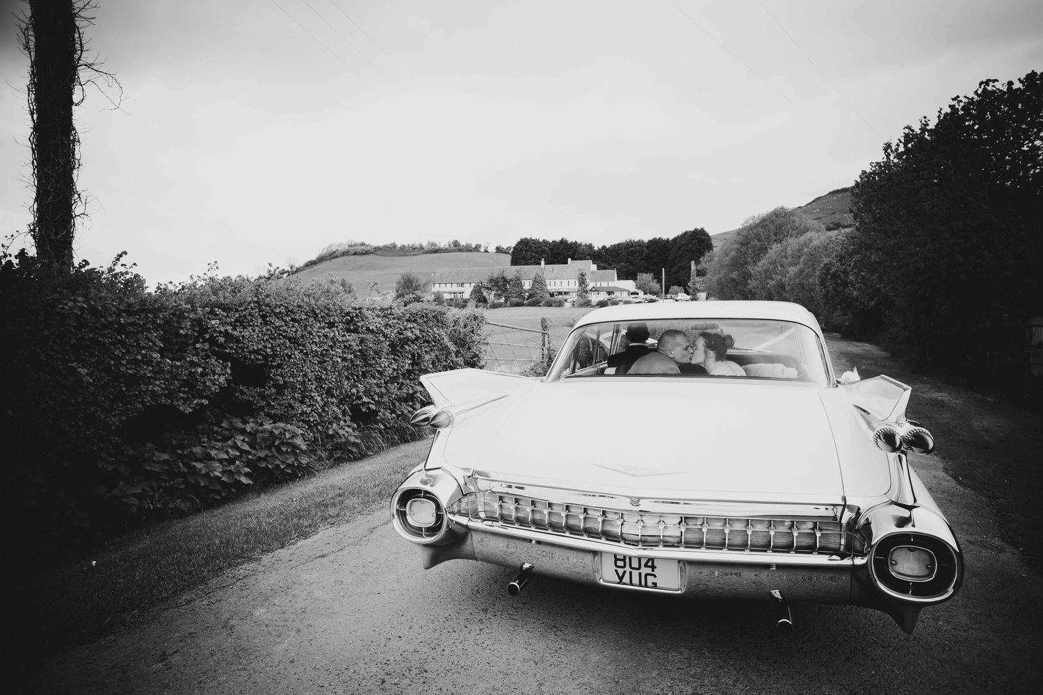 A bride and groom kiss on the back seat of a large 1960's Cadillac parked on a country road