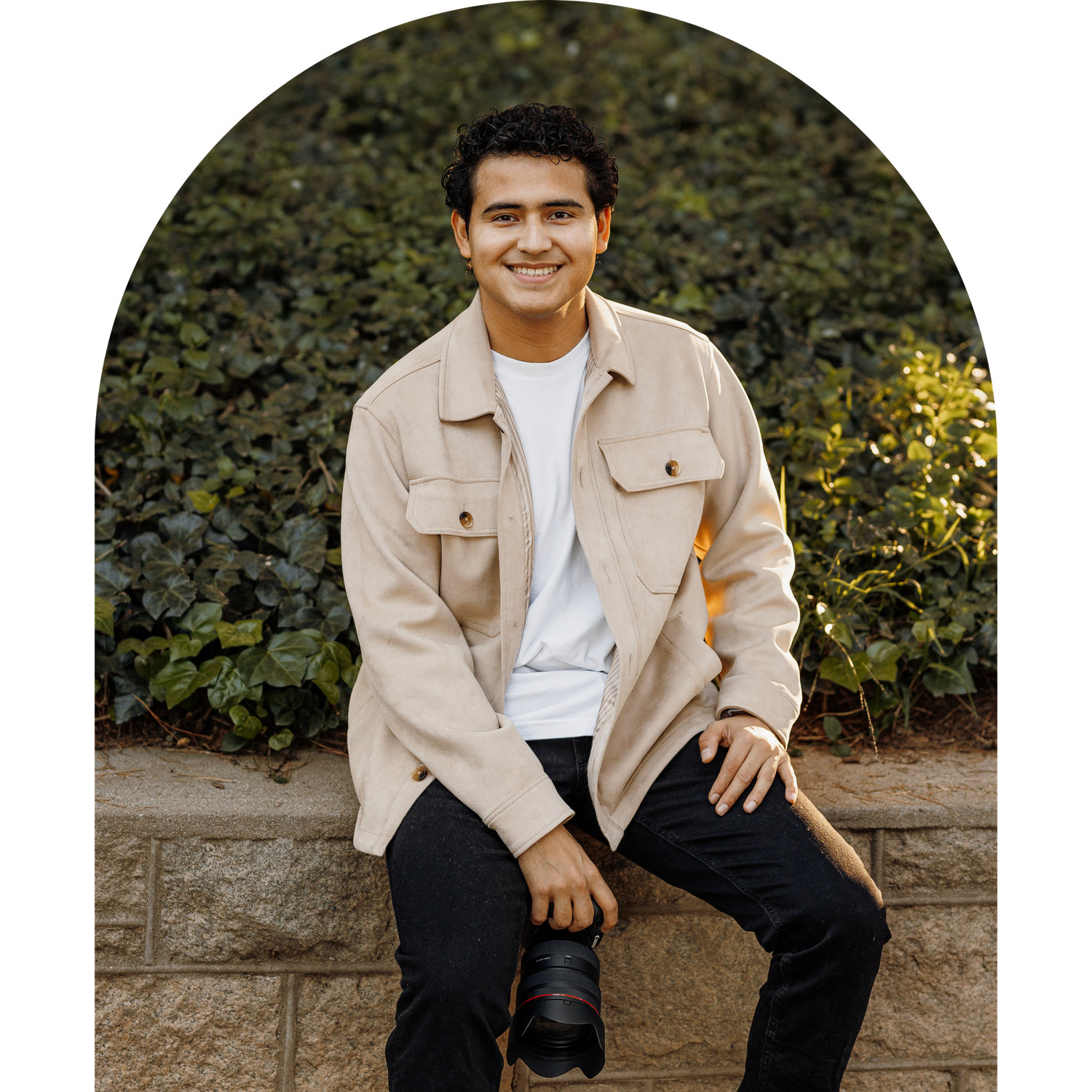 Young man smiling, sitting on a stone wall holding a camera, wearing a beige jacket and black pants.