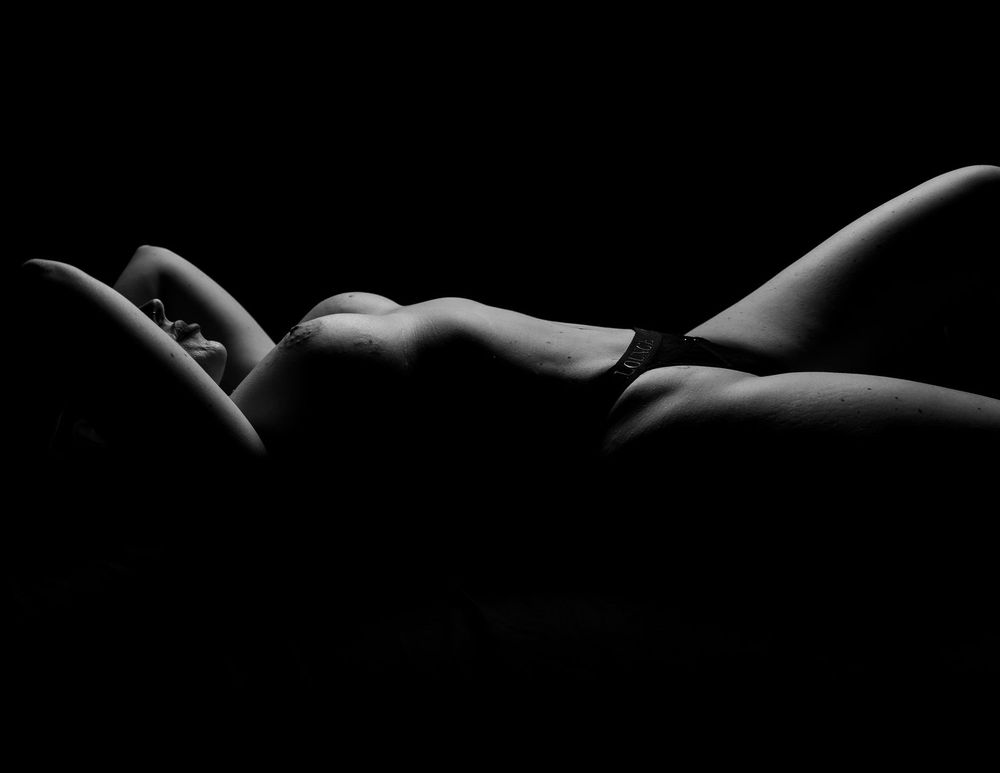 Artistic black and white nude photograph showing shadows and curves of a reclining figure against dark background.