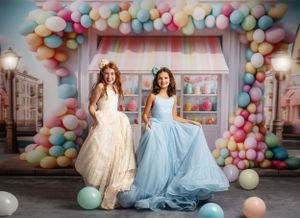 Two flowing dresses in cream and blue twirl against a whimsical candy shop backdrop.