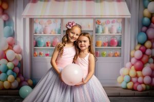 Two dancers in tulle dresses pose with pink balloon against candy shop backdrop with colorful decorations.