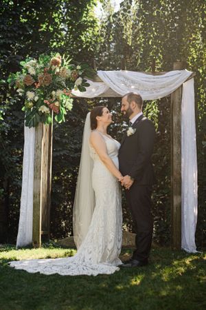 Outdoor wedding ceremony under white fabric arch with lush floral arrangement and greenery backdrop.