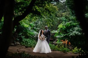 A couple poses on a forest path surrounded by lush green foliage and flowers.