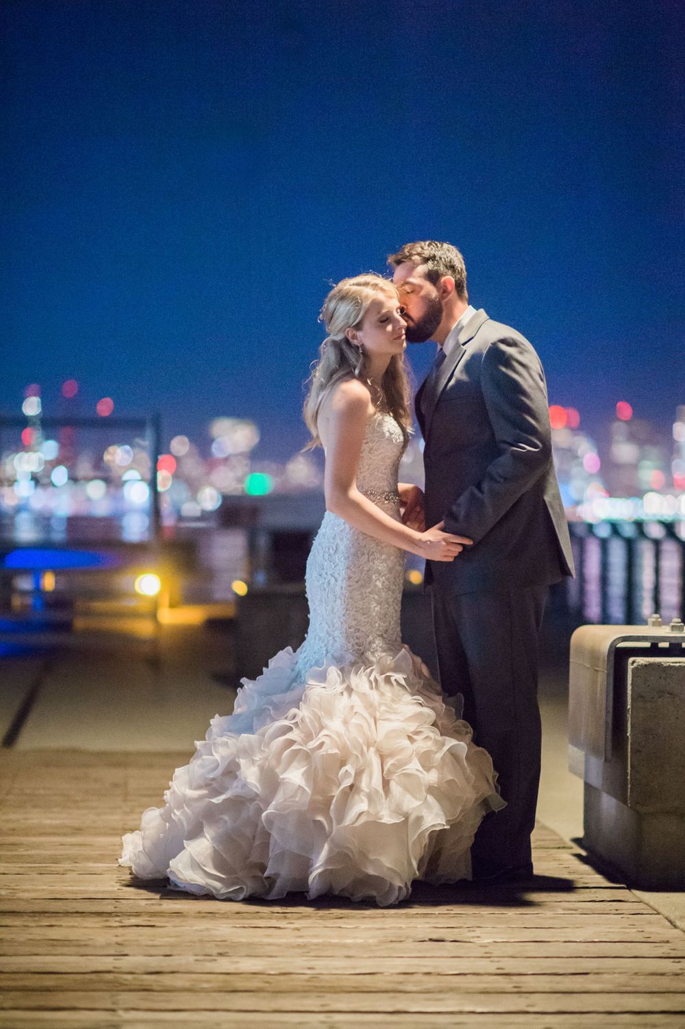A romantic nighttime wedding photo of a couple embracing on a pier with city lights twinkling in the background.
