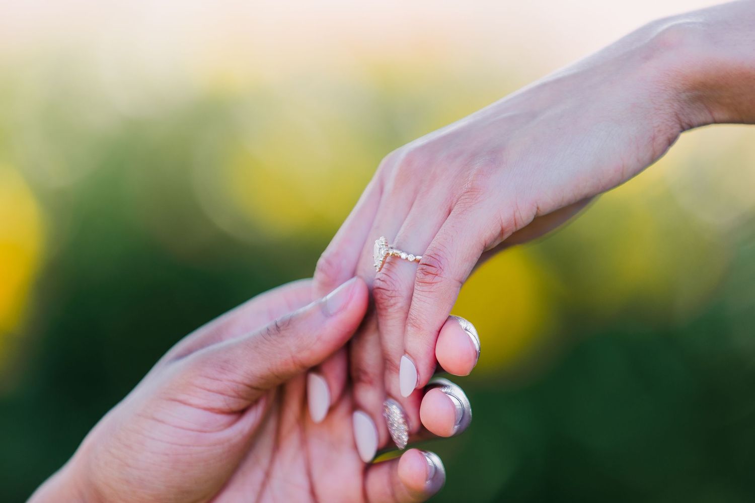 Close-up of two hands touching, one adorned with a delicate diamond engagement ring, against a soft bokeh background.
