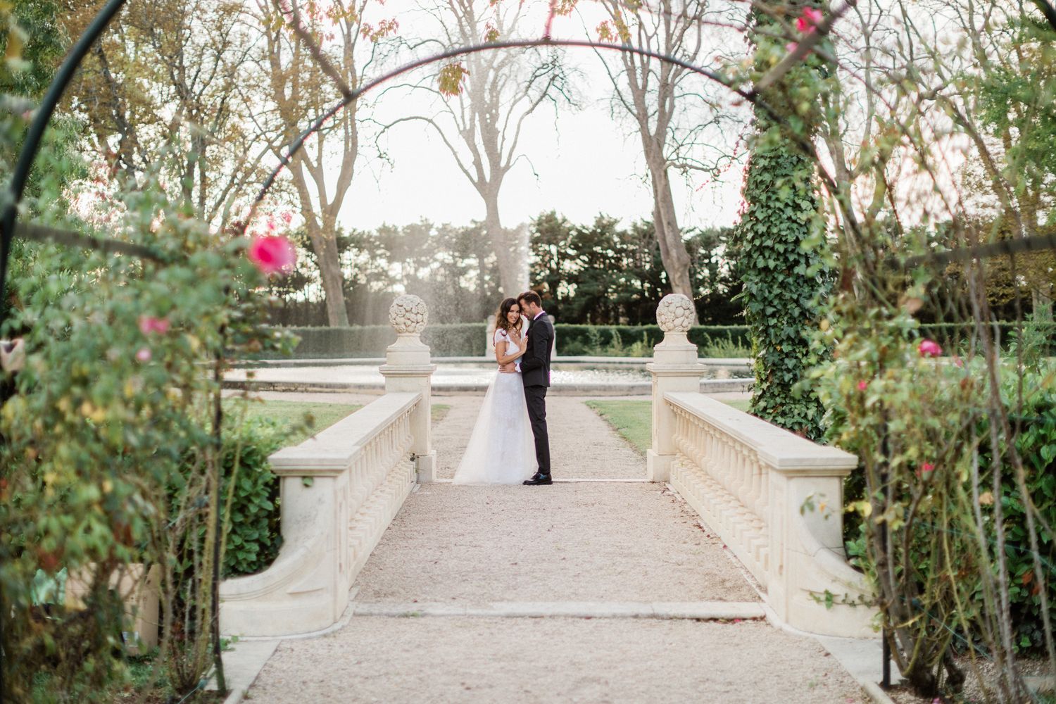 Scène romantique de mariage dans un jardin, avec le couple sur un pont, entouré de vignes fleuries et d’arbres.