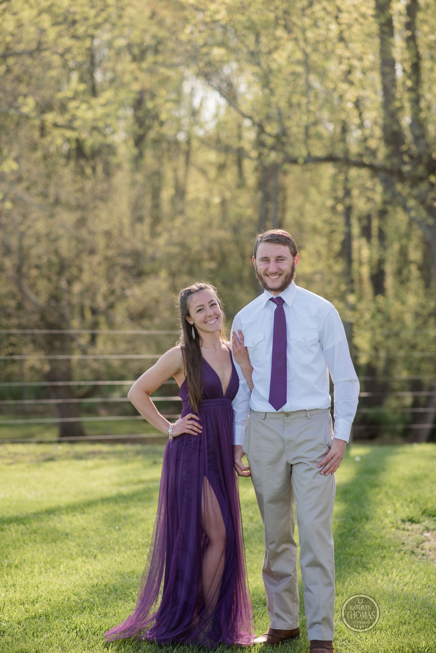 Couple poses on lawn with autumn trees, wearing purple gown and business attire.