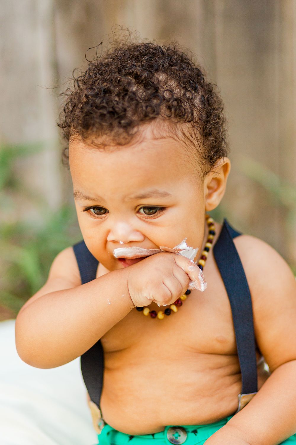 Laughing toddler wearing navy suspenders and turquoise shorts with a beaded necklace during a photoshoot.