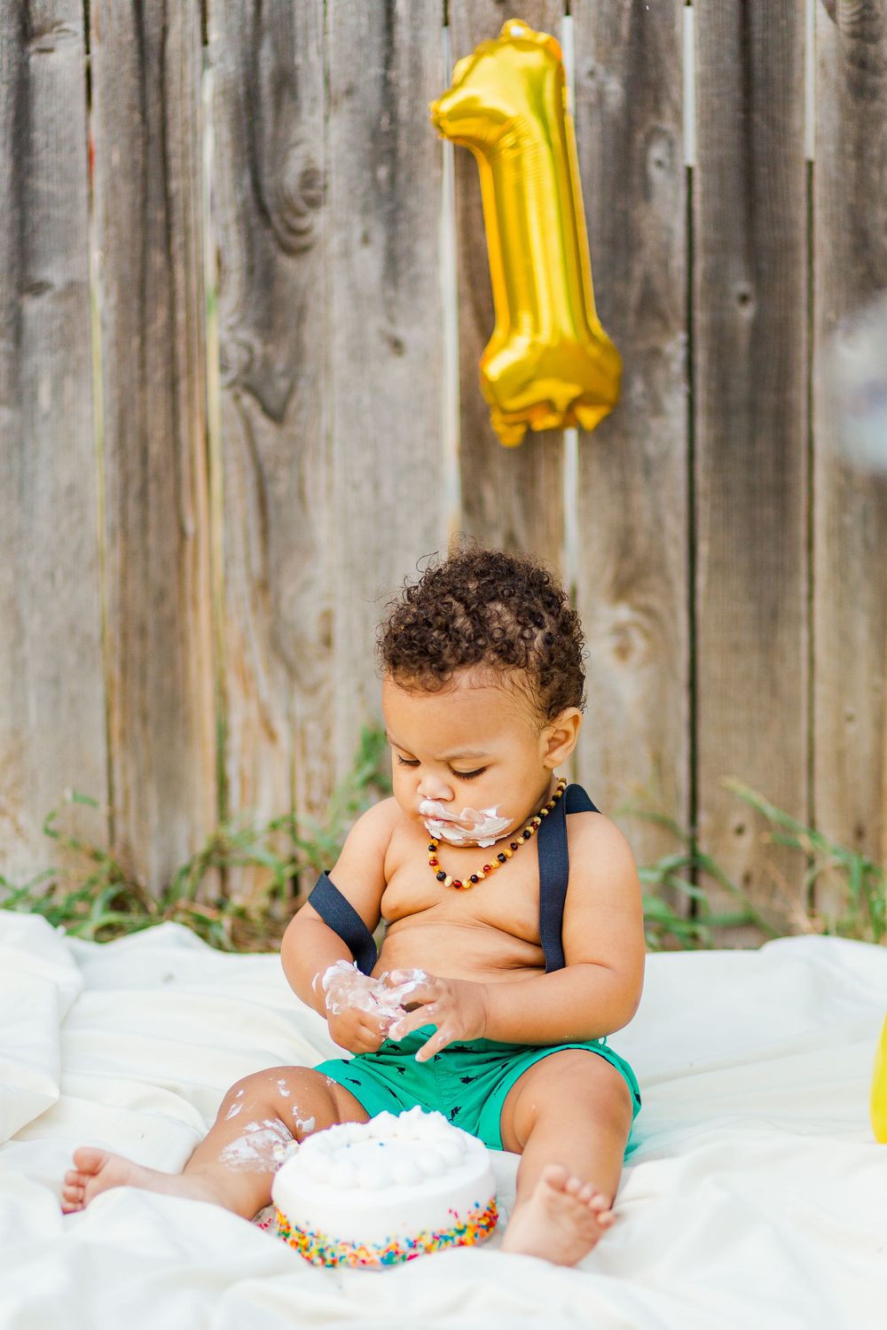 Baby in turquoise shorts plays with smash cake beneath gold number one balloon.