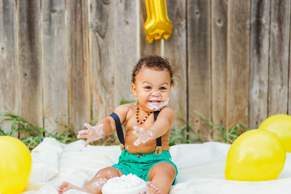 Smiling baby in green shorts enjoys cake smash photo session with yellow balloons.