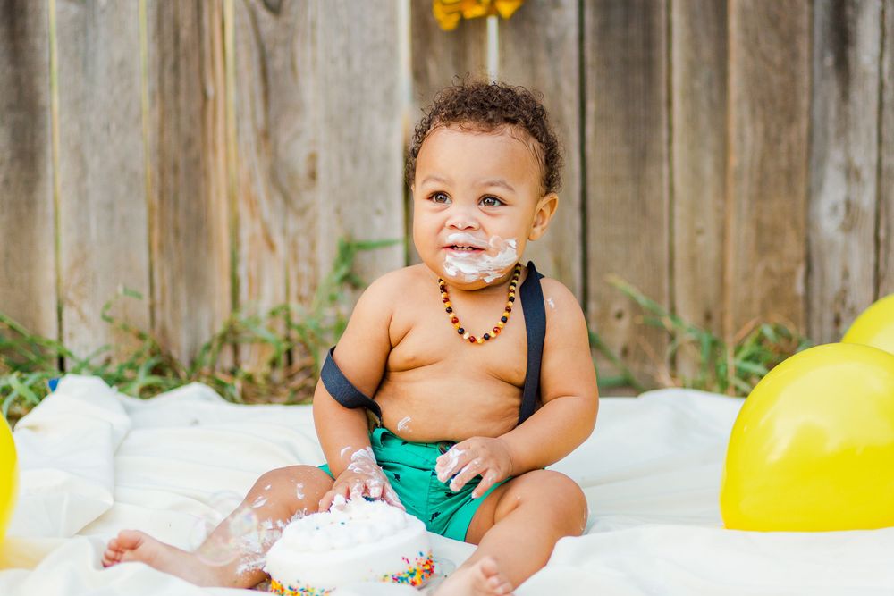 Baby in green shorts sits on white blanket with yellow balloons during cake smash photo session.