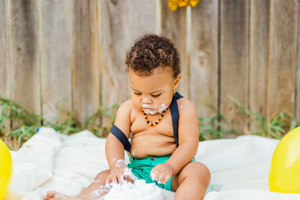 Baby in turquoise shorts plays with cake on white blanket during first birthday photoshoot.