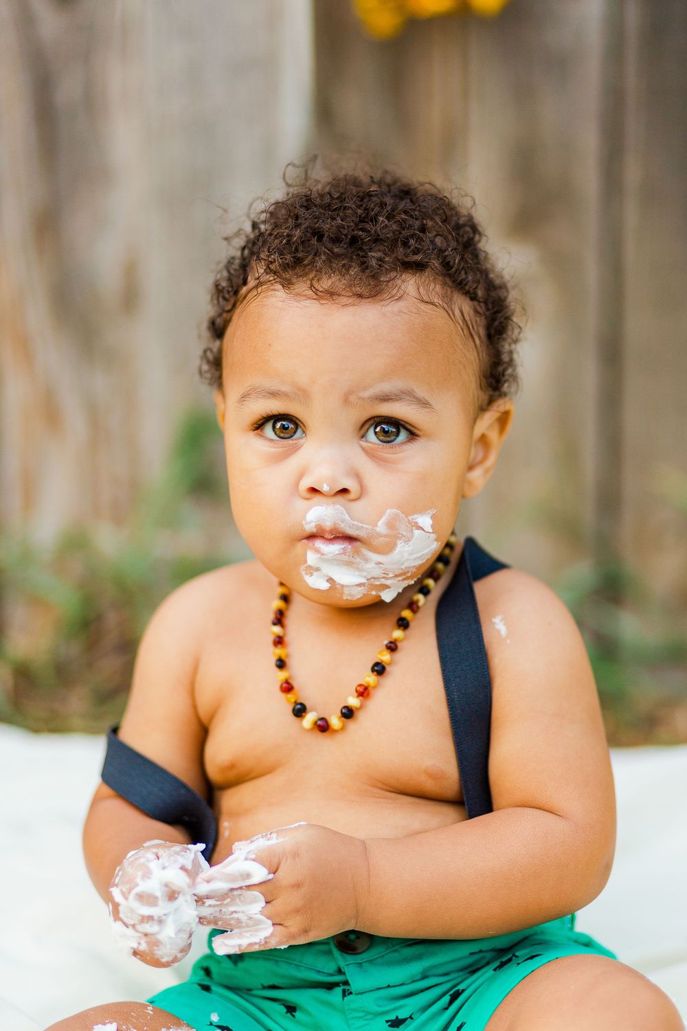 Baby wearing beaded necklace and green shorts has frosting on face during cake smash session.