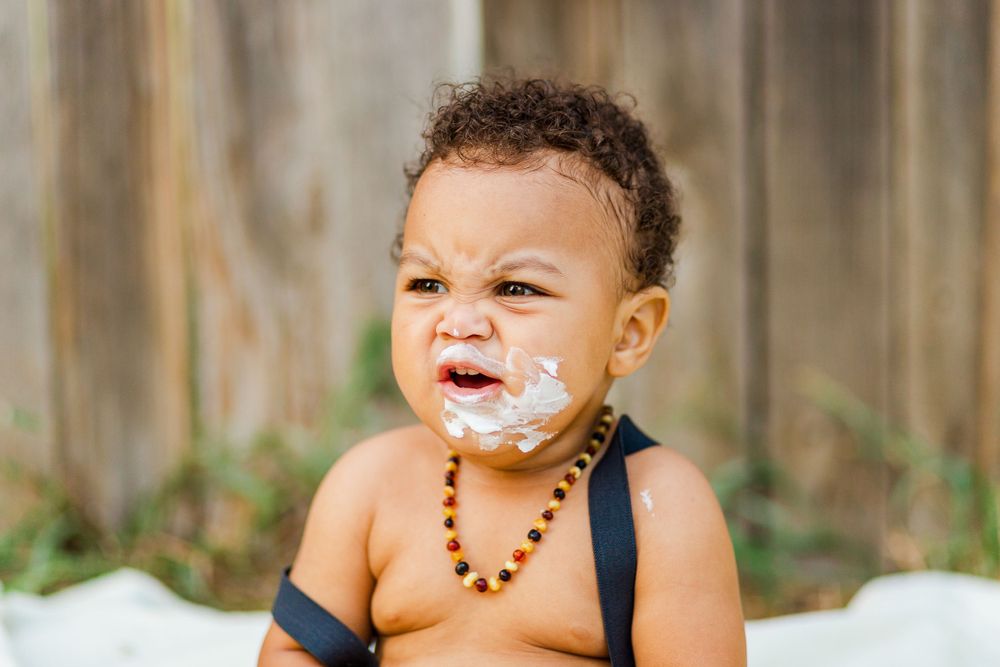 Baby in green shorts and beaded necklace has frosting smeared on face during cake smash.