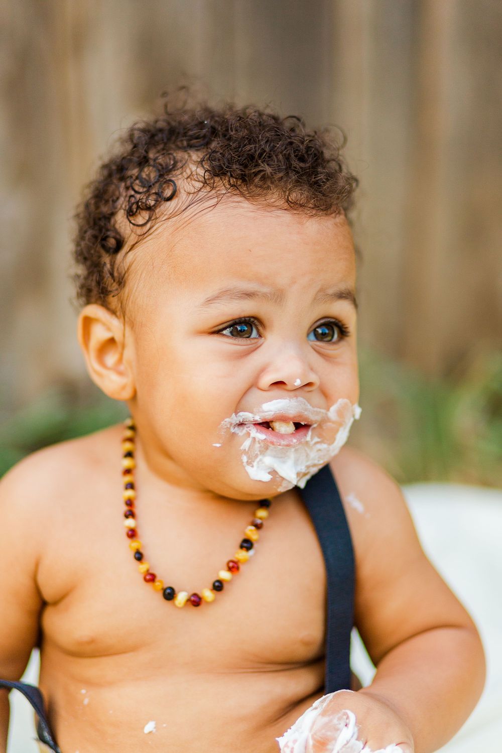 Toddler with curly hair wearing beaded necklace smiles with cake frosting on face.