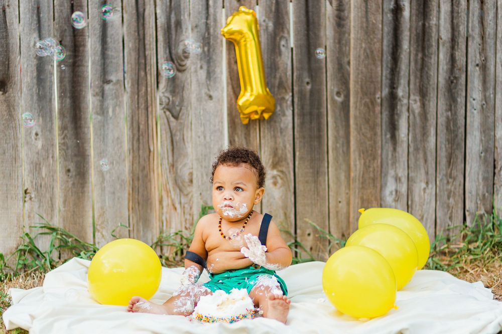 Toddler in turquoise shorts sits on white blanket with yellow balloons and gold number 1 decoration.