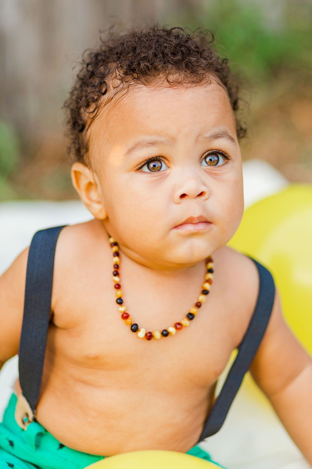 Close-up portrait of toddler wearing beaded necklace and navy suspenders against natural background.