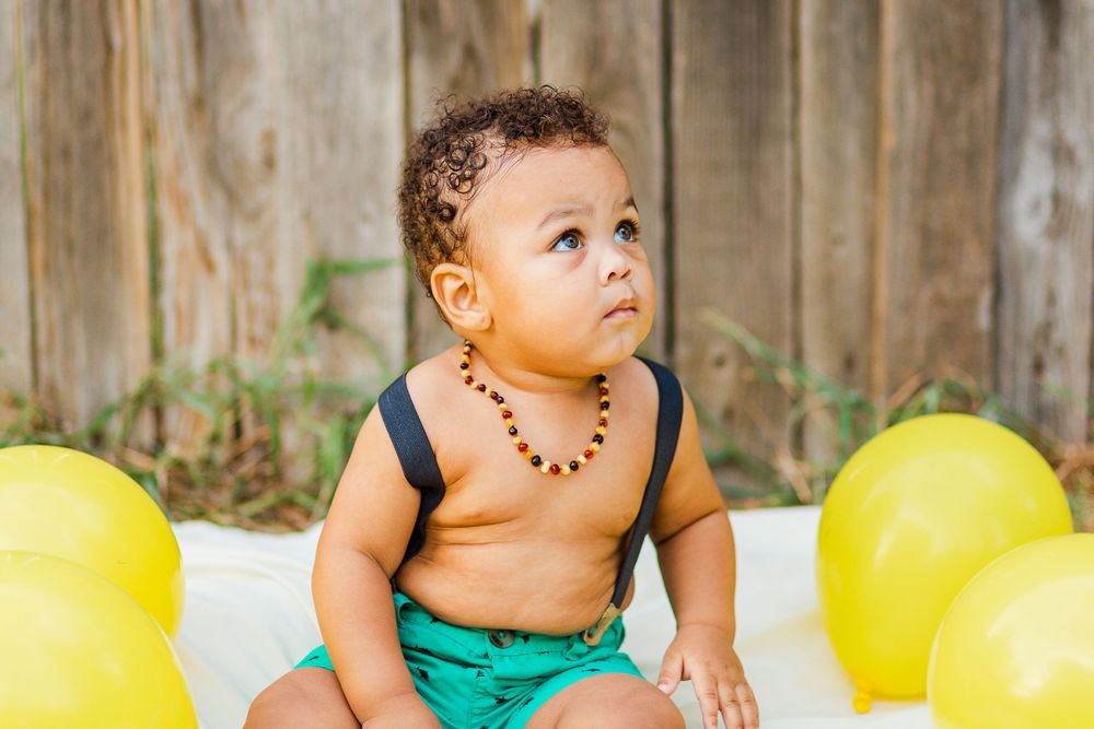 Cute toddler in green shorts and beaded necklace with yellow balloons outdoors.