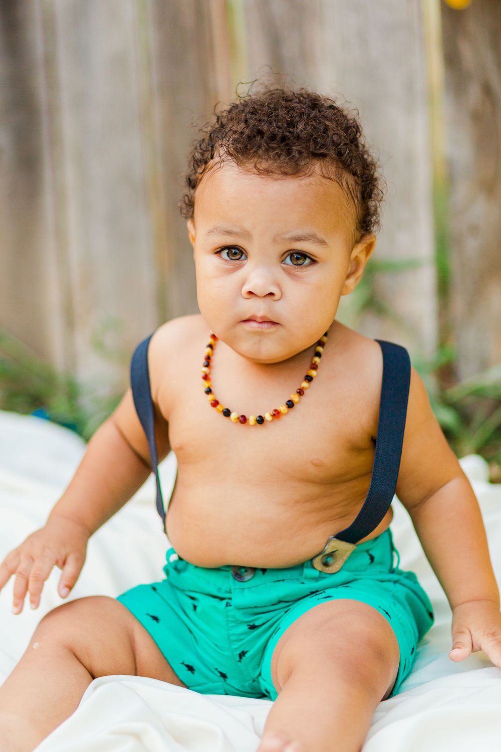 Sweet toddler in turquoise shorts and navy suspenders sitting on white blanket for portrait.