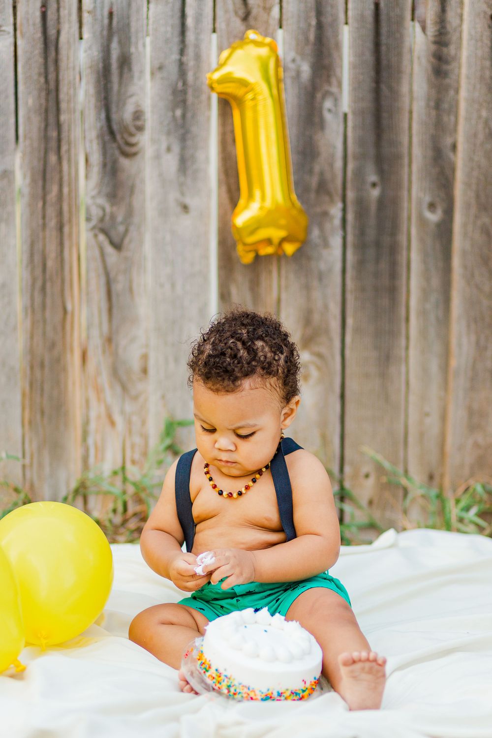 First birthday celebration with cake, yellow balloons and gold number one balloon against wooden fence.