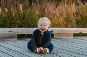 Baby in blue outfit sitting on wooden deck with joyful expression.