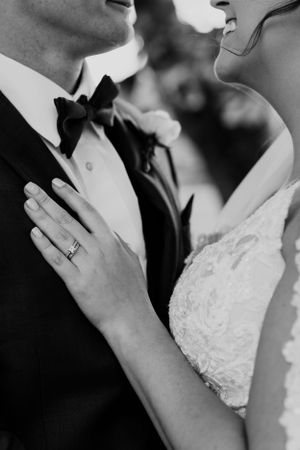 Close-up black and white photo showing bride's hand on groom's tuxedo with wedding ring detail.