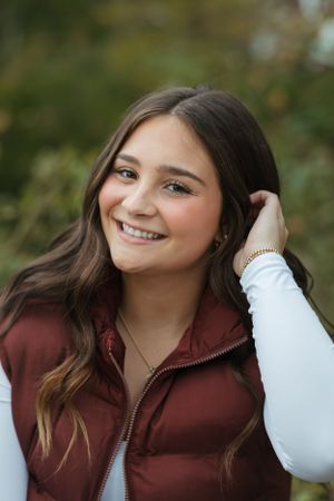 A woman in a burgundy vest and white sweater smiles while touching her hair outdoors.