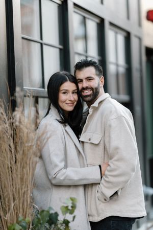 A couple in matching beige coats embraces against a modern building exterior.