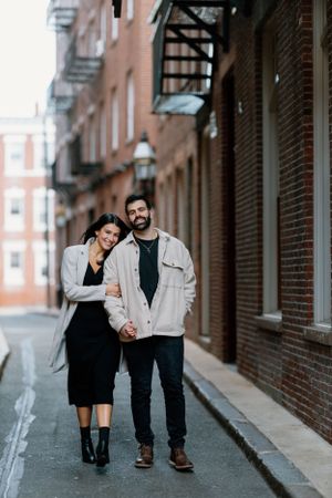A couple poses together on a narrow city street with brick buildings and fire escapes.